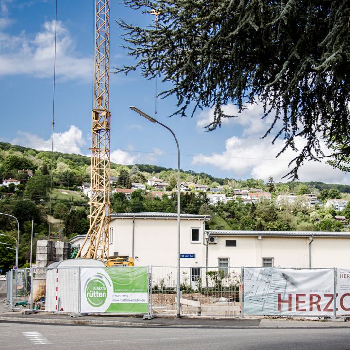 Eine Baustelle mit Werbebannern, Bauzaun und Kran vor einer grünen Hügellandschaft.