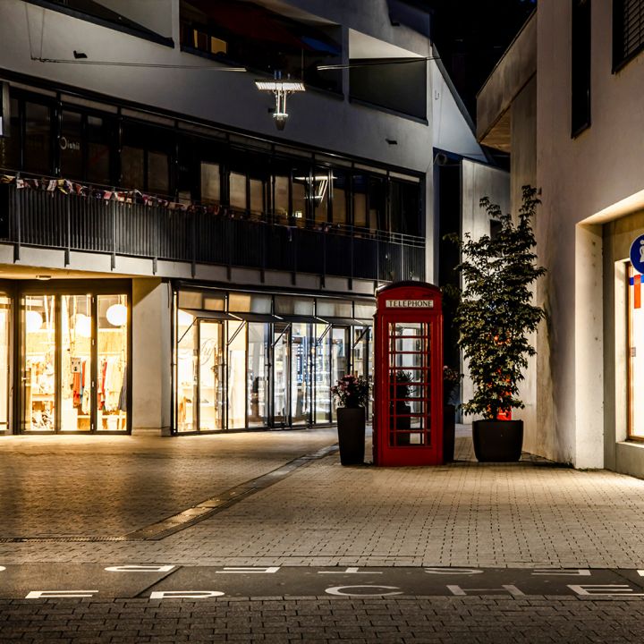Szene einer beleuchteten Fußgängerzone bei Nacht mit einer roten Telefonzelle.