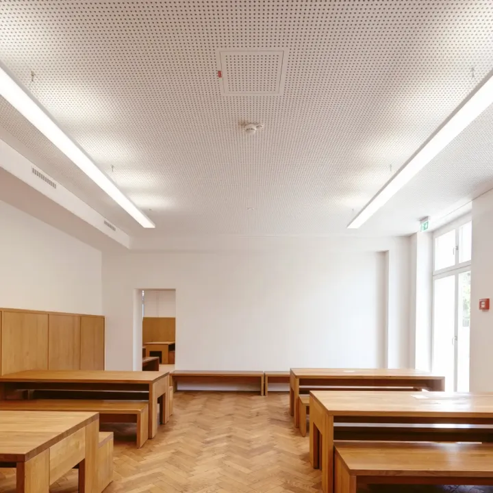 Eine Mensa mit schlichten Holzbänken und moderner Deckenbeleuchtung.
