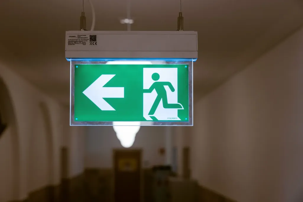Das Bild zeigt ein beleuchtetes grünes Fluchtwegschild mit einem Pfeil nach links und einer rennenden Person zum Ausgang.