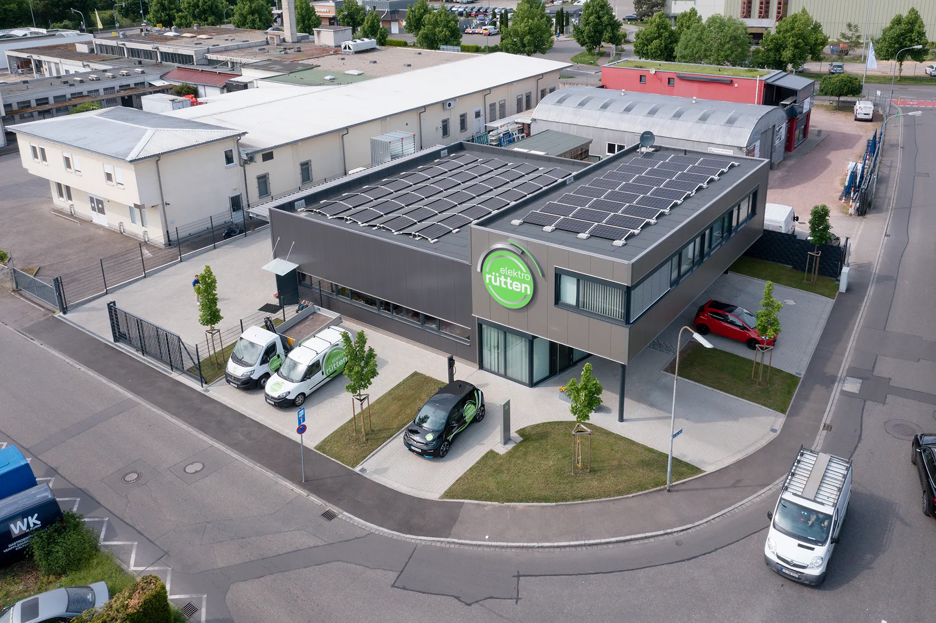 Das Firmengebäude von „elektro rütten“ mit Solaranlage auf dem Dach und Firmenfahrzeugen vor dem Eingang, aufgenommen aus der Vogelperspektive.