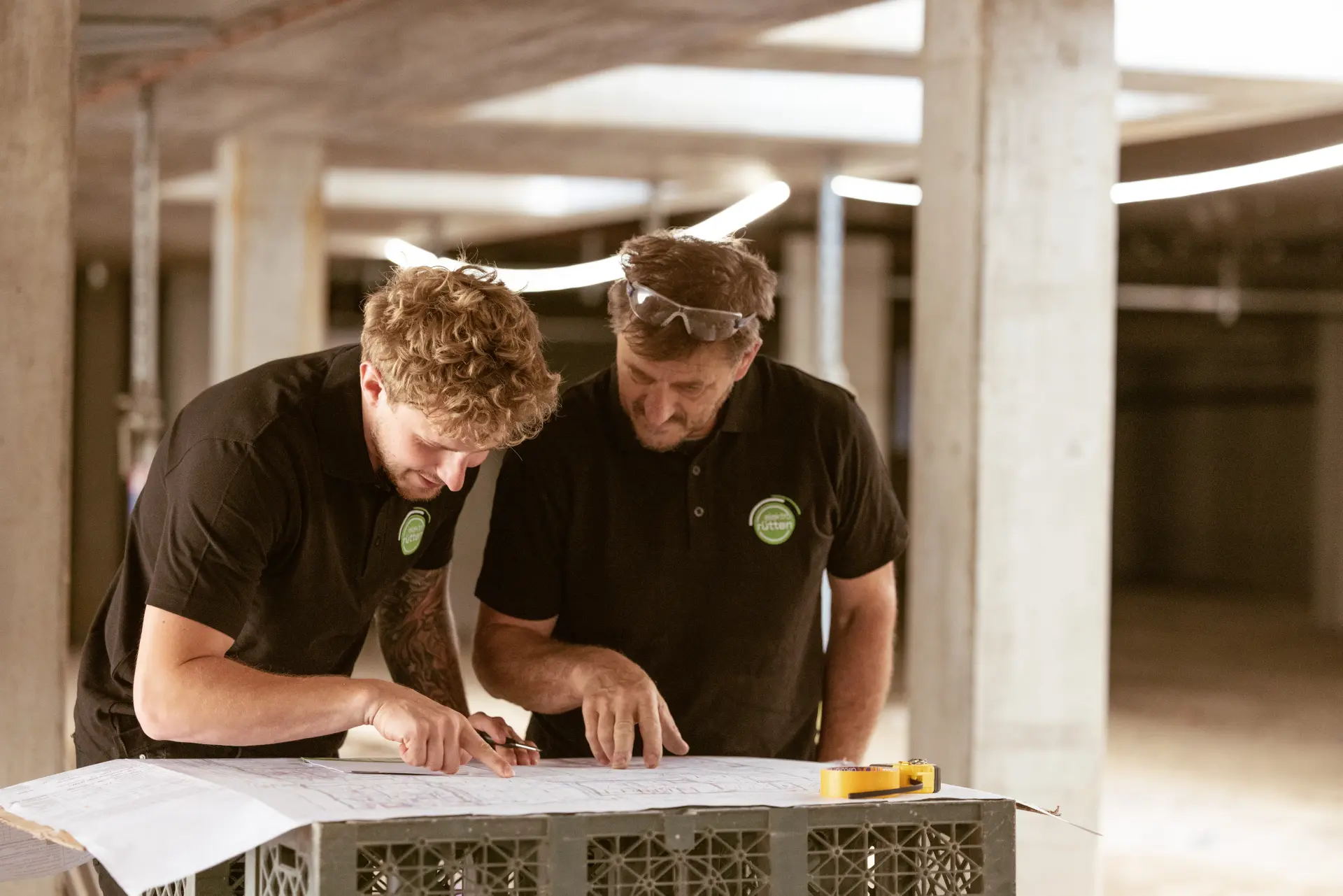 Zwei Mitarbeiter studieren gemeinsam Baupläne auf einer Baustelle.